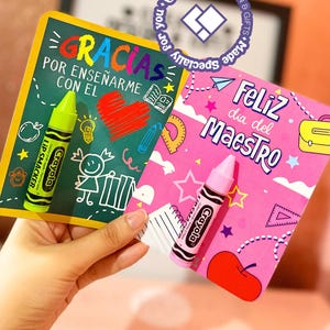 Puede incluir: Dos tarjetas de felicitación con bálsamos labiales Crayola Lip Smacker. Una tarjeta es verde con el texto "GRACIAS POR ENSEÑARME CON EL" y la otra es rosa con "Feliz dia del Maestro". Los bálsamos labiales son amarillos y rosas.