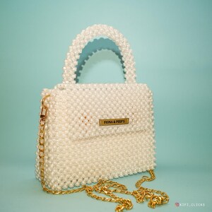 Può includere: Borsa a mano bianca con perline, con manico superiore e tracolla a catena dorata. La borsa presenta una chiusura con patta con una piccola etichetta rettangolare dorata con la scritta "Fiona & Peep's". La borsa è ambientata su uno sfondo azzurro.