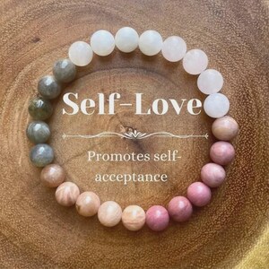 May include: A beaded bracelet with a gradient of colors, from white to pink and peach, with gray-green beads on one side. The text "Self-Love" and "Promotes self-acceptance" are displayed in the center.