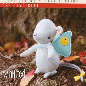 May include: A light blue felt mouse with a yellow button on its wing, sitting on a bed of brown leaves. The mouse is named "Winifred" and is part of the "May blossom designs by Simone Gooding CREATIVE CARD" collection.
