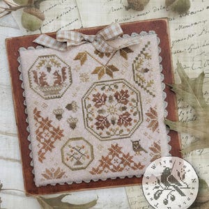 May include: A cross-stitch sampler entitled "Quaker Snippets No. 1" by Brenda Gervais. The design features autumnal motifs like leaves, acorns, and geometric patterns in shades of brown, beige, and green. The sampler is displayed on a wooden surface with a gingham ribbon.