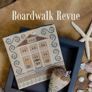 May include: A cross-stitch pattern of a house with a blue and white border, a brown and white pincushion, and the text "Boardwalk Revue" and "Erica Michaels Needleart Designs".