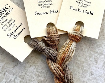May include: Three skeins of embroidery floss in shades of brown, beige, and gold. The floss is labeled "Classic Colorworks" with the color names "Stepping Stones", "Straw Hat", and "Fool's Gold". Each skein is 5 yards of 100% cotton.