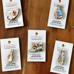 May include: Nine different Peter Rabbit themed needle minders. Each needle minder is a small wooden cutout of a character from the Peter Rabbit stories. The minders are displayed on white cards with the text "Handcrafted Needle Minder" and the website address "thecottageneedles.com".