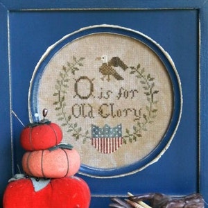 May include: A cross-stitch sampler in a blue frame, featuring the text "O is for Old Glory" and a patriotic design. In front are three tomato-shaped pin cushions. The image also includes the text "Heartstring Samplery" and "The Alphabet Series #1".