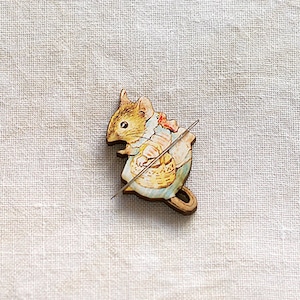 May include: A wooden pin featuring a cartoon squirrel holding a basket and a needle. The squirrel is light brown with a cream-colored belly and a red bow. The pin is set against a neutral linen background.