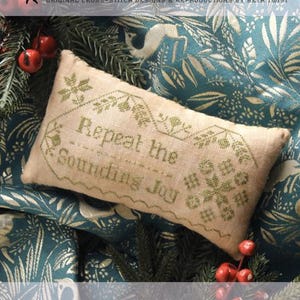Può includere: Un cuscino rettangolare a punto croce con la scritta "Repeat the Sounding Joy" in filo verde. Il cuscino beige è appoggiato su un tessuto verde acqua con un motivo floreale. L'immagine include il testo "Heartstring Samplery".