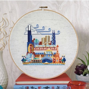May include: A cross-stitch embroidery hoop with a colourful Chicago skyline design. The design includes iconic buildings, a Ferris wheel, and the text "pretty little chicago". The hoop is displayed on a stack of books.