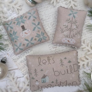 May include: Three embroidered cushions with winter designs. One shows a snowman with a top hat, another a Christmas tree, and the third says "let's build snowmen". The cushions are beige with blue and white details.