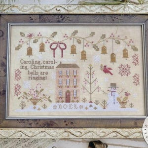 以下が含まれることがあります： クリスマスのテーマの額入りクロスステッチアートワーク。家、雪だるま、鐘が描かれており、「Caroling, caroling, Christmas bells are ringing!」というテキストが特徴です。フレームは明るいベージュ色です。