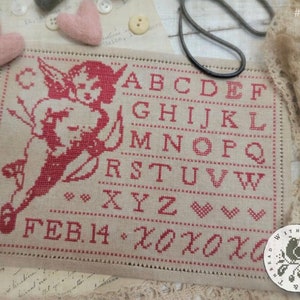 May include: A cross-stitch sampler with a red and white design of a cupid holding an arrow and a heart. The sampler also includes the alphabet, the date February 14th, and the phrase "XOXOXO".