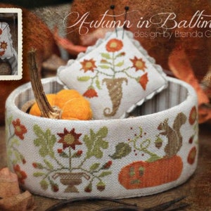 May include: A cross-stitch design featuring a decorative basket with a pumpkin and a small pillow. The basket is adorned with floral and squirrel motifs in orange, green, and brown. The text "Autumn in Baltimore" is visible.