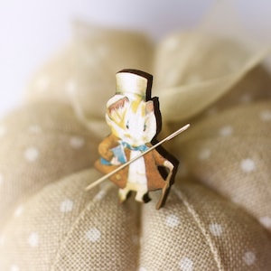 Puede incluir: Un cojín de alfileres de tela beige con lunares blancos y una figurita de madera de un ratón con un sombrero de copa y un palo.
