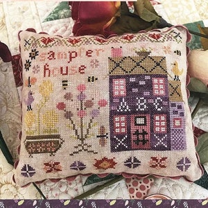 Puede incluir: Un sampler de punto de cruz con una casa en tonos rosa y morado. El sampler se titula "Sampler House" y forma parte de la serie "Houses on Wisteria Lane Series". El diseño es de Lori Pengelly.