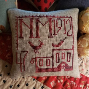 May include: A small, beige pillow with red embroidery depicting a house with the number "1912" above it. The house has a chimney and a bird perched on the roof.