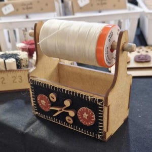 May include: Wooden thread holder with a spool of white thread and a black fabric base with orange and white floral designs. The holder has a small compartment for storing sewing supplies.