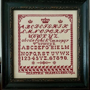 May include: A framed cross-stitch sampler with a red alphabet, numbers, and crowns. The sampler is on a white background and has a border of red leaves. The text at the bottom reads "MARTHA MASIALER 57894".
