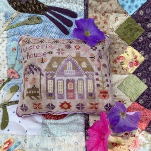 May include: A cross-stitch pillow with a purple house and the text "Wisteria House" on a white background. The pillow is surrounded by purple and pink flowers on a patchwork quilt.