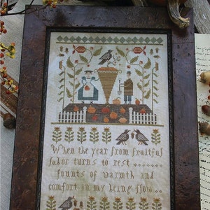 Puede incluir: Un bordado de punto de cruz enmarcado con un tema otoñal. El diseño presenta una cesta de fruta, dos figuras y pájaros. El texto dice "Cuando el año de un trabajo fructífero se vuelve al descanso... manantiales de calidez y consuelo fluyen en mi ser..."