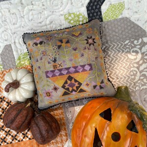 May include: A cross-stitch pillow with a Halloween theme featuring a purple and yellow pot with black bats, orange pumpkins, and a black cat. The pillow is on a white and orange patchwork quilt with a jack-o-lantern and gourds.