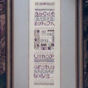 May include: A framed cross-stitch sampler with a white background and purple and gray embroidery. The sampler features the alphabet, numbers, and various decorative designs.