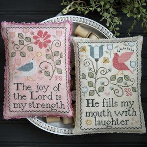 May include: Two decorative cross-stitch pillows with floral and bird designs. One pillow has the text "The joy of the Lord is my strength." The other says "He fills my mouth with laughter."