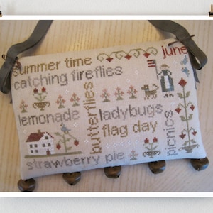 May include: A beige fabric pillow with a stitched design featuring summer-themed words and images, including "summer time", "catching fireflies", "lemonade", "butterflies", "ladybugs", "flag day", "strawberry pie", and "picnic". The pillow has a brown leather strap and brown bells attached to the bottom.