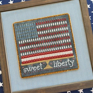 Puede incluir: Obra de arte bordada en punto de cruz enmarcada que representa la bandera estadounidense. El diseño presenta rayas rojas y blancas, estrellas azules y las palabras "sweet liberty". La obra está enmarcada en un marco de madera.