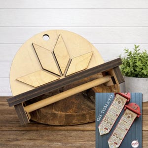 May include: A wooden paper towel holder with a natural wood finish and dark brown accents. The holder features a circular top with diamond-shaped cutouts and a horizontal bar for the paper towels. A small, framed cross-stitch pattern with the text "OH TOMATO" is also visible.
