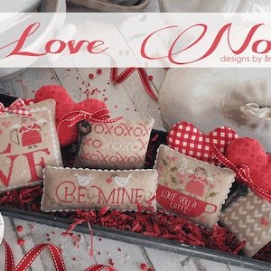 May include: A collection of Valentine's Day themed cross-stitch designs. The image features several small, embroidered pillows with phrases like "LOVE" and "BE MINE", along with heart-shaped decorations. The color palette includes red, beige, and white.
