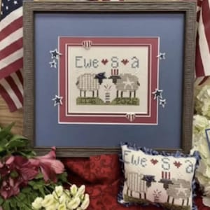 May include: Framed cross-stitch artwork with a patriotic theme. The design features two sheep with the text "Ewe S a" and the years "2026" and "2026". The artwork is framed in a brown wooden frame and surrounded by blue and red accents.