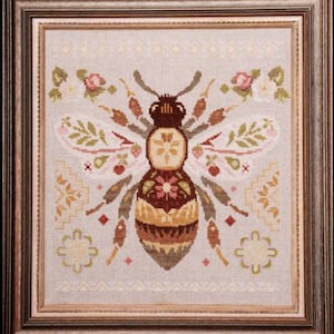May include: Cross-stitch artwork in a wooden frame, depicting a detailed bee design. The bee is in shades of brown, beige, and gold, with floral and leaf accents. The background is light beige. The frame is made of wood.