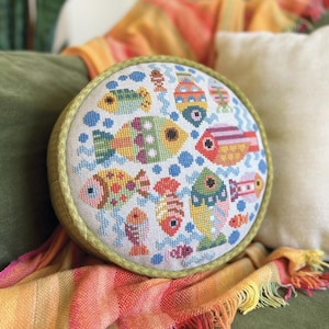 May include: Round decorative pillow with a colorful cross-stitch fish pattern. The pillow features various fish designs in shades of yellow, green, orange, and pink, with blue accents. The pillow has a light green border and is placed on a green surface.
