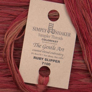 Può includere: Un'etichetta marrone con il testo "SIMPLY SHAKER Sampler Threads COLORFAST by industry standards The Gentle Art counted thread embroidery PO. Box 670 New Albany, Ohio 43054 RUBY SLIPPER 7100" e un piccolo foro nell'angolo in alto a destra. L'etichetta è circondata da un mucchio di filo da ricamo rosso.