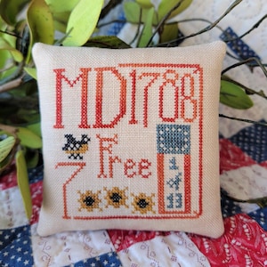 Może przedstawiać: Mała, biała poduszka z haftem w kolorze czerwonym, niebieskim i żółtym. Poduszka zawiera napis "Made Free" i małą flagę amerykańską.