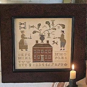 May include: A framed cross-stitch sampler depicting a house, figures, and floral designs in muted tones. The artwork includes letters, numbers, and decorative motifs. The frame is dark brown with a textured finish, adding to the vintage aesthetic.