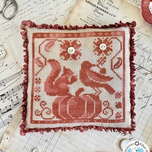 May include: A cross-stitch pillow with a red and white design of two birds perched on a pumpkin. The pillow has a ruffled edge and is surrounded by vintage paper and buttons.