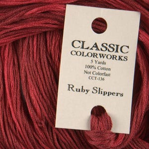 Può includere: Un gomitolo di filo da ricamo rosso etichettato "Classic Colorworks" con il nome del colore "Ruby Slippers" e i dettagli "5 Yards, 100% Cotone, Non Colorfast, CCT-136".