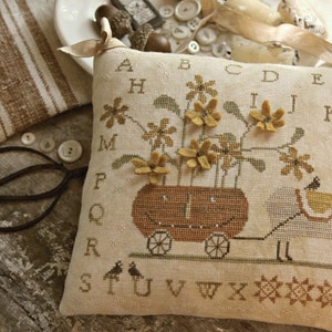 Puede incluir: Un cojín decorativo beige con un diseño de punto de cruz. El cojín presenta una oveja tirando de un carro lleno de flores. El diseño incluye el alfabeto y el texto "BOO & BAA-BBIE design by Brenda Gervais".