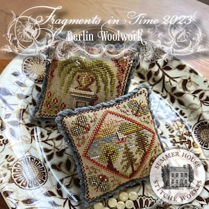 May include: Two small, hand-stitched needlepoint pillows with a gray trim. The pillows are on a white and brown patterned plate. The pillows feature floral and landscape designs. The text "Fragments in Time 2023" and "Berlin Woolwork" are visible above the pillows. The text "Summer House Stitch Works" is visible below the pillows.