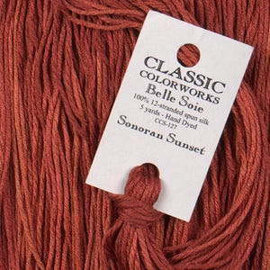 May include: Close-up of a skein of Classic Colorworks Belle Soie thread in the color "Sonoran Sunset." The label indicates it is 100% 12-stranded spun silk, hand-dyed, and 5 yards in length.