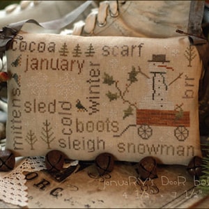 May include: A beige cross-stitch pillow with a snowman design and the words "cocoa", "scarf", "january", "mittens", "sled", "winter", "boots", "sleigh", and "snowman". The pillow is decorated with a brown ribbon and a metal clasp. The pillow is sitting on a table with a book and a lace doily.