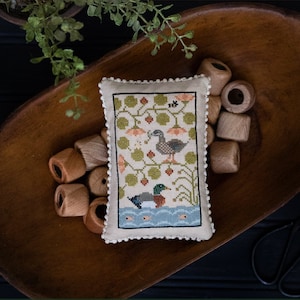 Può includere: Un cuscino ricamato a punto croce con un'anatra marrone e bianca e un'anatra verde in uno stagno con foglie verdi e fiori arancioni. Il cuscino è in una ciotola di legno con bobine di filo di legno. Il testo "Quack Stack" è sotto l'immagine. Il testo "Original Designs by Paulette Stewart" è sotto il testo "Quack Stack".