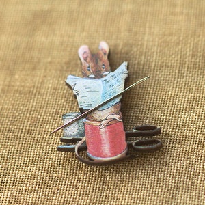 Op de afbeelding: Een grillige broche met een cartoonmuis die een krant leest met de titel "The Tailor and Cutter". De muis bevindt zich boven een rode garenklos, met een naald en een schaar. De broche is geplaatst tegen een bruin jute achtergrond.