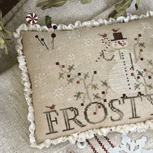 Puede incluir: Un cojín beige bordado con un diseño de muñeco de nieve y la palabra "Frosty". El muñeco de nieve lleva un sombrero de copa y una bufanda. El cojín tiene un borde con volantes y está decorado con copos de nieve y pájaros.