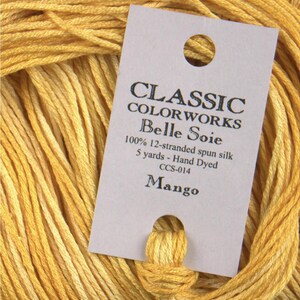 May include: Close-up of a skein of yellow-orange Classic Colorworks Belle Soie thread. The label reads "Mango" and indicates it is 100% 12-stranded spun silk, 5 yards, hand dyed. The thread is a vibrant, warm color, ideal for embroidery or needlework projects.