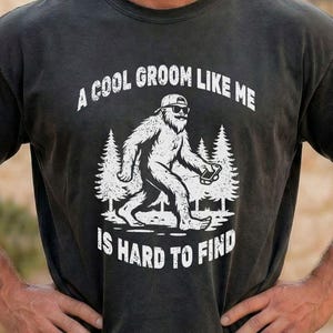 Op de afbeelding: Zwart t-shirt met een witte afbeelding van een Bigfoot-achtig wezen met een zonnebril en een telefoon. De tekst luidt: "A COOL GROOM LIKE ME IS HARD TO FIND".