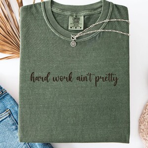 May include: An olive green t-shirt with the phrase "hard work ain't pretty" in a script font. The shirt is folded and accessorized with a silver necklace, blue jeans, and white sneakers. A dried palm leaf adds a natural touch.