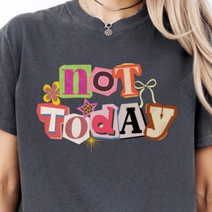 May include: Dark gray t-shirt with the words "NOT TODAY" in a colorful, cut-out style design. The text is accented with floral and star graphics. The shirt has short sleeves and a relaxed fit.