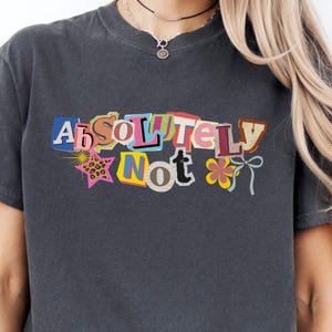 May include: Dark gray t-shirt with the words "Absolutely Not" in a colorful, cut-out style design. The design includes a star, a flower, and a bow. The shirt is a casual, unisex style.